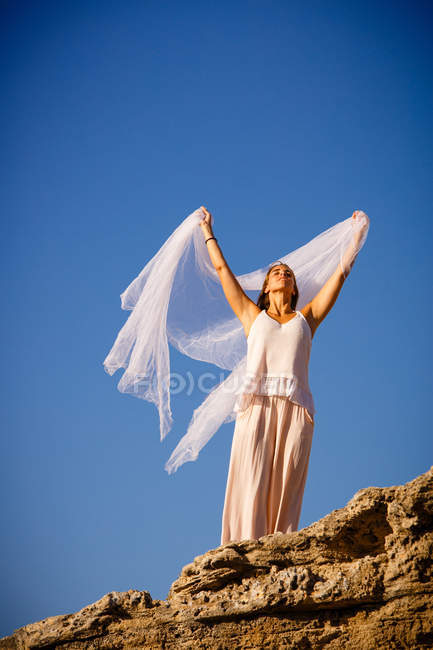 Von unten junge geheimnisvolle Frau mit erhobenen Händen, die weißes Textil hält und auf Felsen und blauem Himmel posiert — Stockfoto