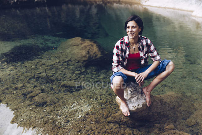 Felice donna adulta seduta sulla roccia in tranquilla acqua trasparente del lago godendo la natura e sorridendo via — Foto stock