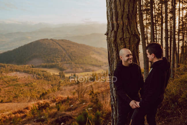 Alegre pareja homosexual mirándose el uno al otro cerca del árbol en el bosque y pintoresca vista del valle - foto de stock