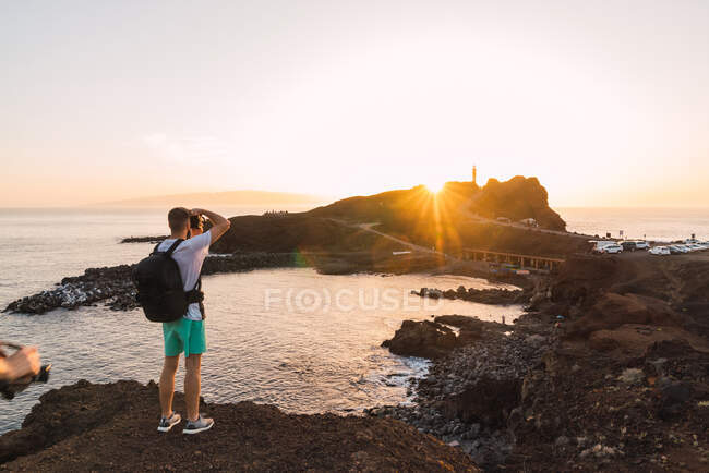 Vue arrière du touriste avec sac à dos tournage à la caméra coucher de soleil sur la côte rocheuse sous forme de baie de mer à Tenerife, Îles Canaries, Espagne — Photo de stock