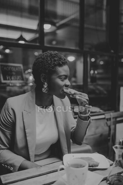 African American elegant woman eating sandwich and sitting at table with cup of drink in street cafe — Stock Photo