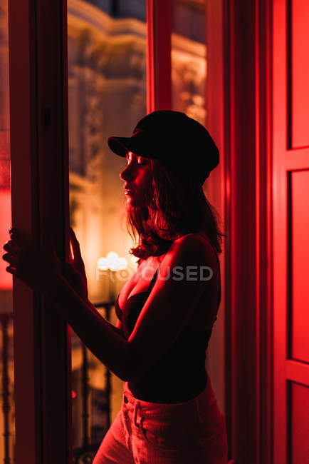 Молода тонка жінка в Кап стоїть біля балкона в кімнаті в почервоніння вночі — стокове фото