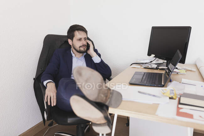 Концентрований молодий чоловік з ногами на столі розмовляє на мобільному телефоні і сидить у кріслі в офісі — стокове фото