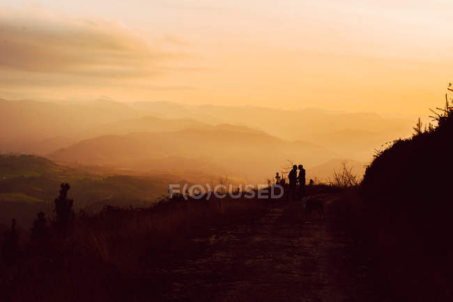 Distante casal homossexual abraçando perto do cão na rota em montanhas ao pôr do sol — Fotografia de Stock