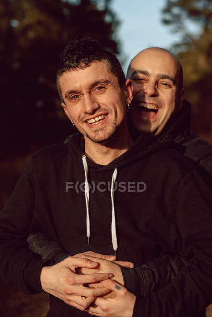 Retrato de alegre pareja homosexual abrazándose en el bosque en un día soleado sobre un fondo borroso - foto de stock