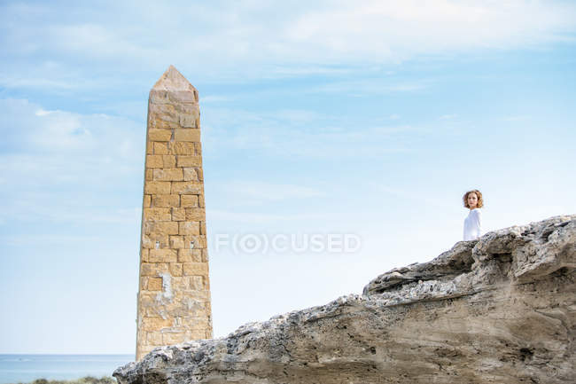 Pensive woman standing near stone construction in form of tower on rocky sea coast — Stock Photo