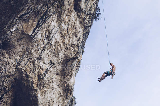 De baixo escalador pendurado na corda em penhasco áspero contra o céu azul — Fotografia de Stock