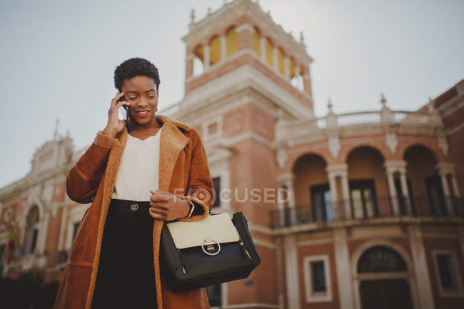 Charming confident African American elegant woman in jacket holding bag and mobile phone on street — Stock Photo