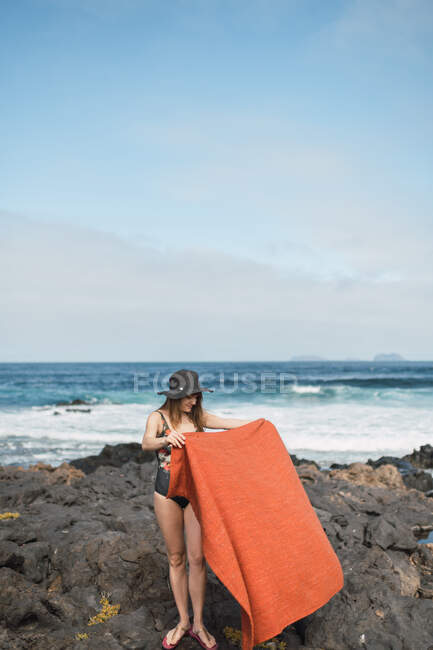 Slim hembra en traje de baño y sombrero que pone la toalla en la costa pedregosa cerca del mar ondulante en la magnífica naturaleza - foto de stock