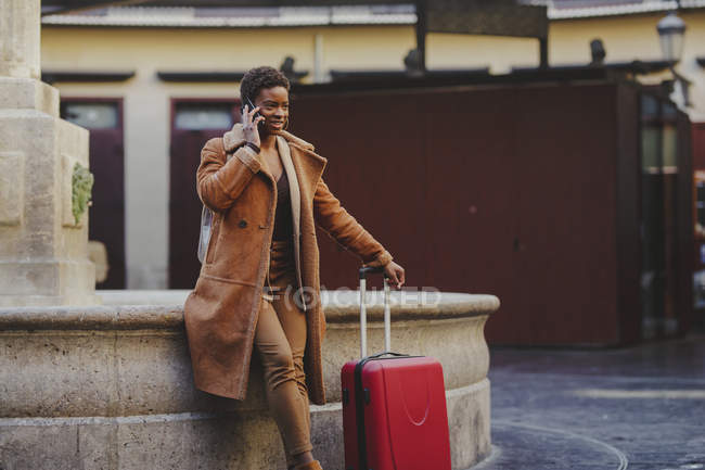 Donna afroamericana elegante con bagaglio parlare sul cellulare e in piedi vicino monumento sulla strada — Foto stock