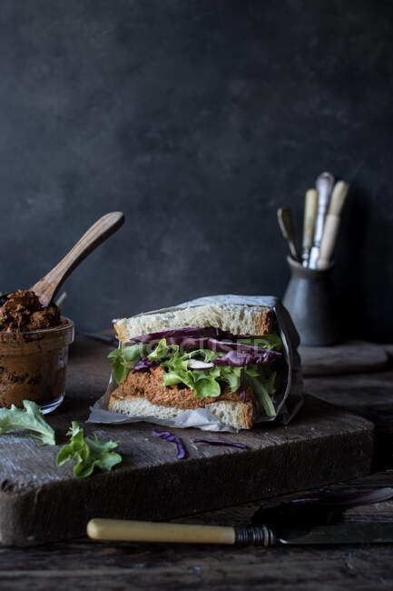 Sandwich aus Pastete mit trockenen Tomaten, frischem Salat und Kohl auf Tablett neben Messer auf Holzbrett auf schwarzem Hintergrund — Stockfoto