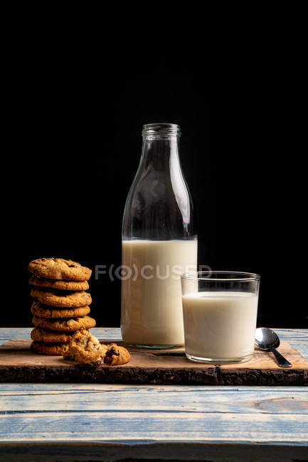 Glass and bottle of milk and pile of fresh cookies on wooden board on black background — Stock Photo