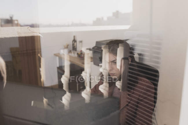 Vue latérale de la jeune femme naviguant sur téléphone portable et assise à table avec ordinateur portable dans le bureau par la fenêtre — Photo de stock