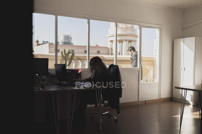 Вид збоку молодої леді, що переглядає ноутбук і сидить за столом в офісі і чоловік розмовляє на мобільному телефоні на балконі біля церкви в сонячний день — стокове фото