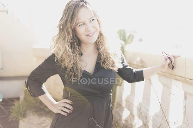 Молодая элегантная веселая леди в платье с рукой на бедре держит очки и позирует на балконе в солнечный день — стоковое фото