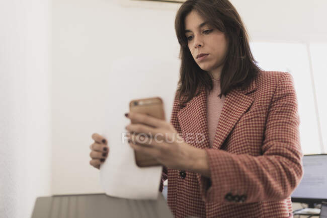 Jeune femme concentrée naviguant sur téléphone portable et imprimant des documents au bureau sur fond blanc — Photo de stock