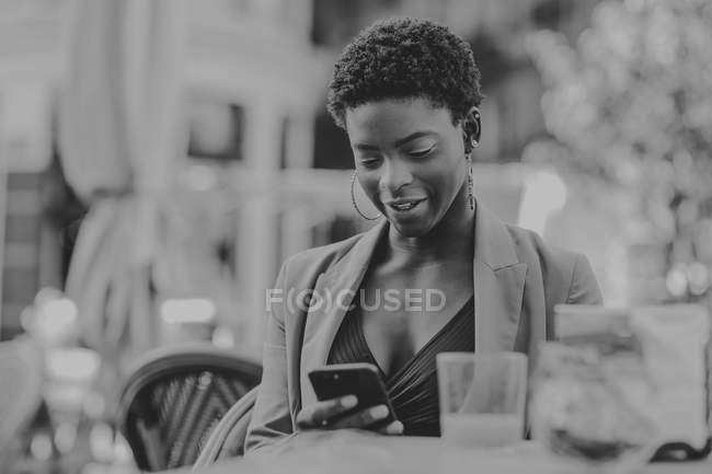 Femme élégante afro-américaine joyeuse tenant un téléphone portable et assise à table avec un verre de jus dans un café de rue — Photo de stock