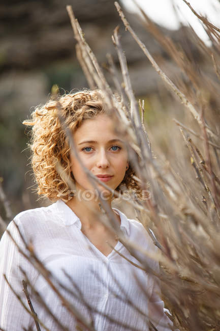 Joven mujer encantadora mirando a la cámara cerca de ramas secas de arbusto sobre fondo borroso - foto de stock