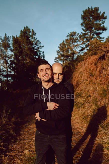 Retrato de alegre casal homossexual abraçando na floresta em dia ensolarado no fundo borrado — Fotografia de Stock
