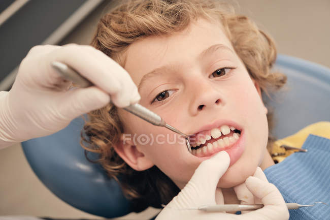 Manos de dentista en guantes utilizando herramientas profesionales para examinar los dientes de lindo niño en la clínica - foto de stock