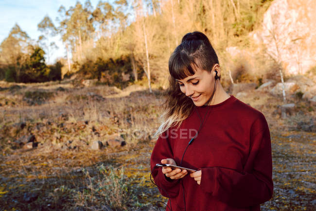 Junge Hipster-Frau mit Piercing und Kopfhörer hört Musik mit Handy und geht auf Landstraße — Stockfoto