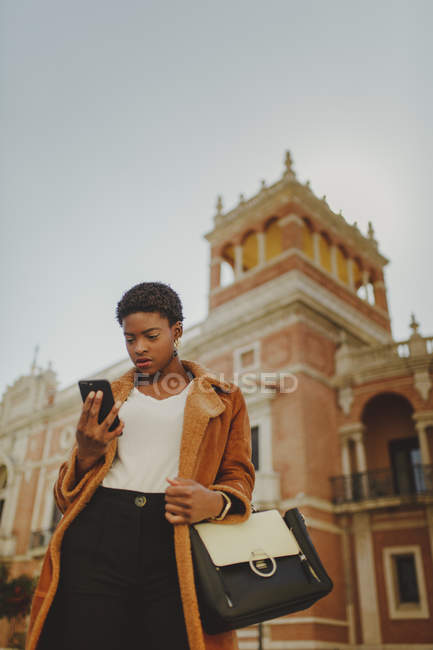 Confident African American elegant woman in jacket holding bag and mobile phone on street — Stock Photo
