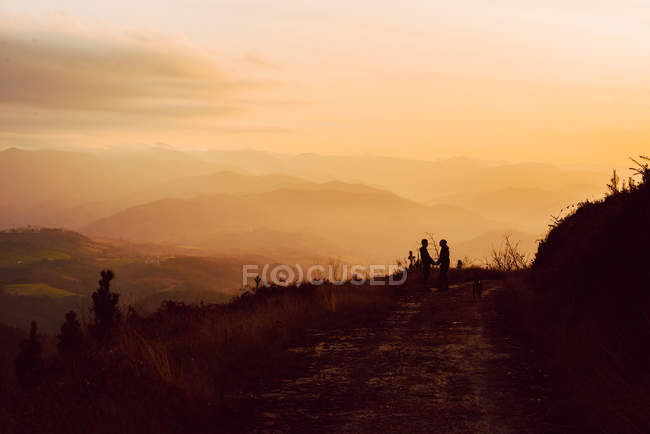 Далека гомосексуальна пара обіймається біля собаки на шляху в гори на заході сонця — стокове фото