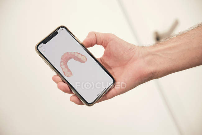 Mano de un hombre irreconocible que demuestra un teléfono inteligente moderno con escaneo de dientes en la oficina del dentista - foto de stock