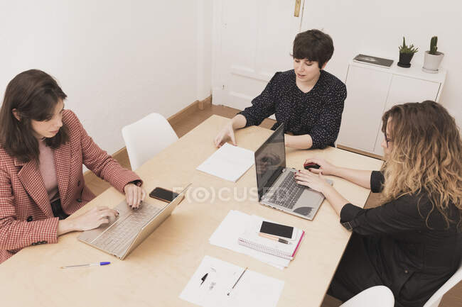 Concentrated young females near lady browsing on mobile phone and typing on laptop at table in office — Stock Photo