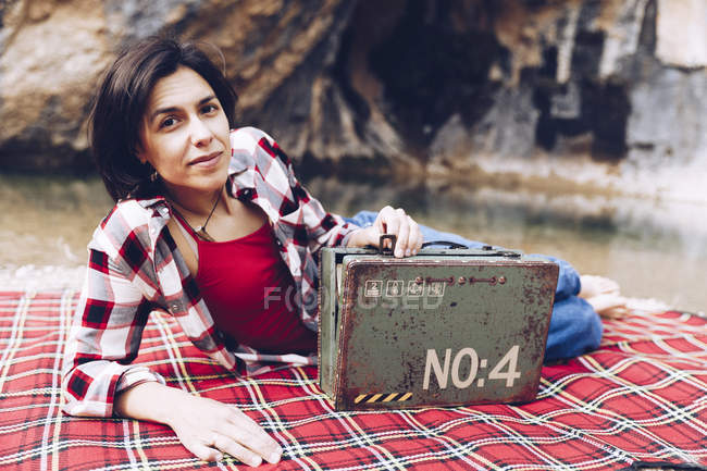 Улыбающаяся взрослая женщина лежит на берегу озера в скалах, держа старый ржавый чехол с провиантом, улыбаясь в камеру — стоковое фото