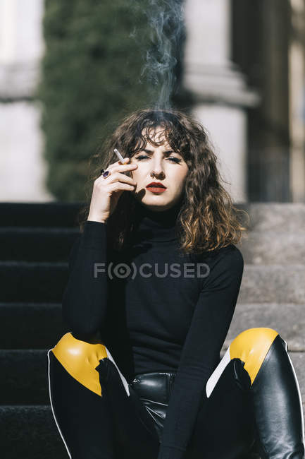 Attraente giovane donna in abito elegante seduta sulle scale e fumare sigarette nella giornata di sole sulla strada della città — Foto stock