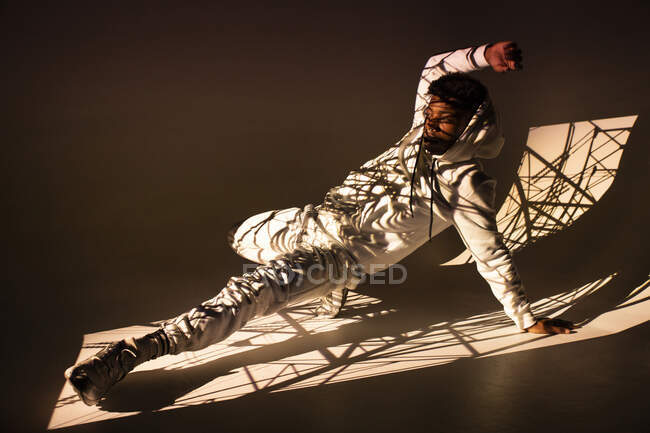 Beau mâle afro-américain en vêtements de sport effectuant un mouvement de danse sous la lumière vive de la fenêtre sur fond brun — Photo de stock