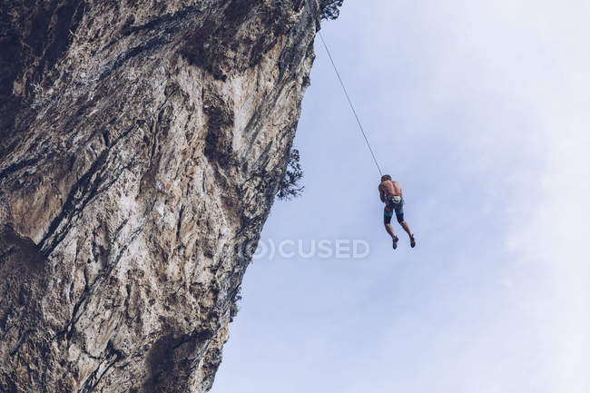 From below unrecognizable climber hanging on rope on rough cliff against blue sky — Stock Photo