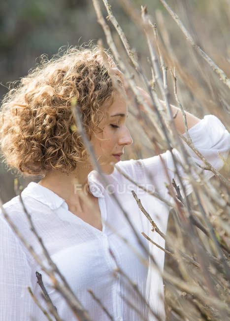 Молодая задумчивая женщина, стоящая возле сухих ветвей кустарника на размытом фоне — стоковое фото