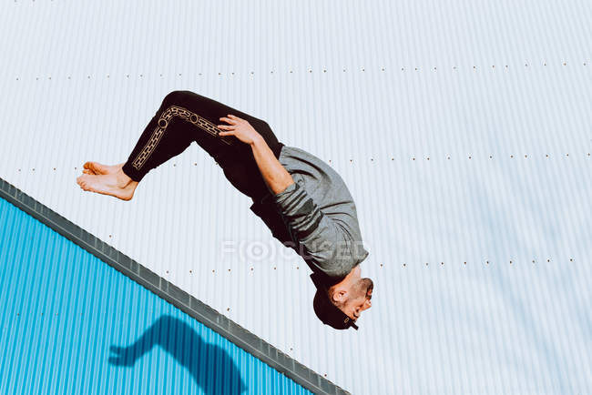Босоногий парень в стильном наряде, выполняющий сальто у стены современного здания — стоковое фото