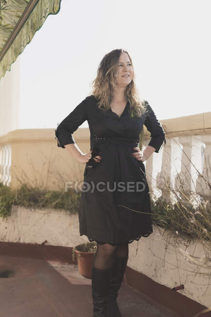 Jeune dame élégante et joyeuse en robe avec la main sur la hanche tenant des lunettes et posant sur le balcon dans la journée ensoleillée — Photo de stock