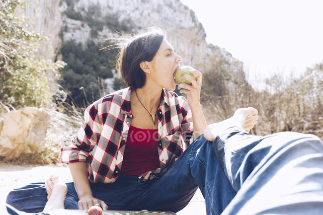 Mann liegt auf Plaid, Frau sitzt daneben und beißt Apfel auf und genießt die gemeinsame Zeit beim Picknick in der Natur — Stockfoto