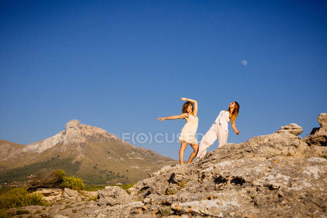Mujeres jóvenes misteriosas con las manos levantadas posando en rocas cerca de la colina y el cielo azul con la luna - foto de stock