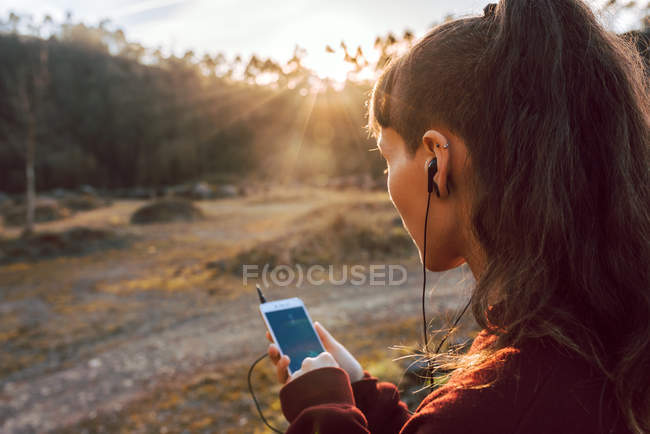 Joven mujer hipster con piercing y auriculares escuchando música con teléfono móvil en el campo - foto de stock