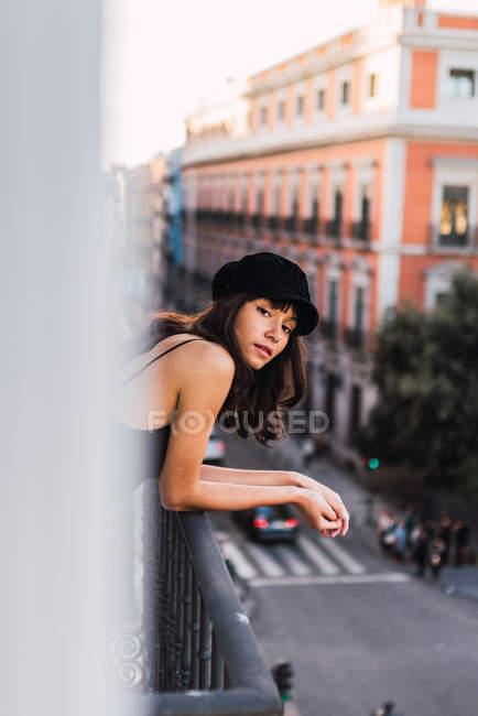 Молода струнка жінка в кепці дивиться на камеру і стоїть на балконі біля вулиці з вогнями ввечері — стокове фото
