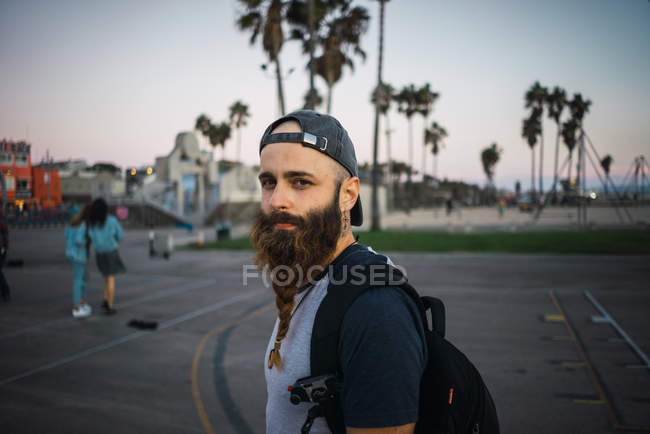 Vista lateral de tipo barbudo con mochila mirando a la cámara mientras está de pie en el pavimento en la calle de la ciudad moderna - foto de stock