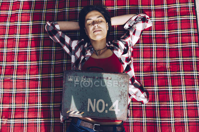 Smiling adult woman lying on plaid on lake shore in cliffs holding old rusty case with provision smiling at camera — Stock Photo