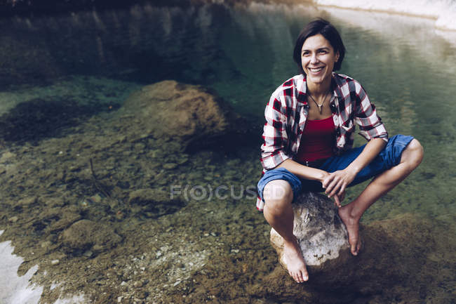 Glückliche erwachsene Frau sitzt auf einem Felsen im ruhigen, transparenten Wasser des Sees und genießt die Natur und lächelt weg — Stockfoto