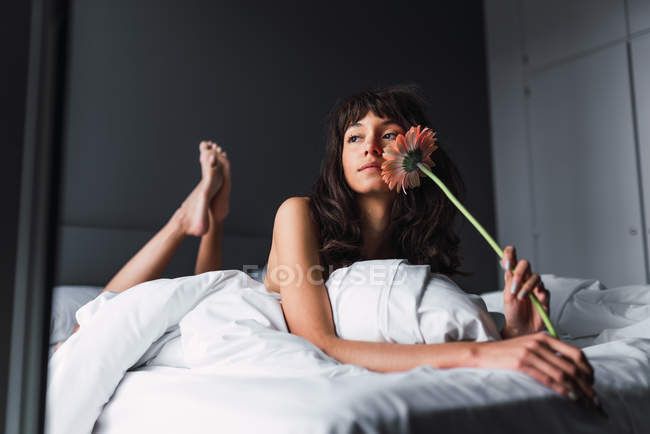 Jovem mulher atraente com flor fresca olhando para longe e descansando na cama no quarto — Fotografia de Stock