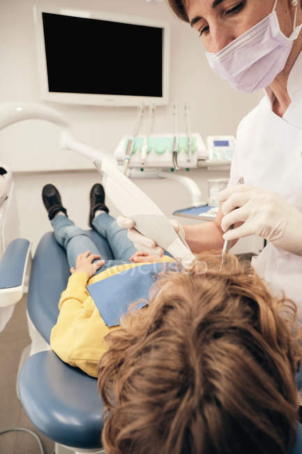 Woman in mask and doctor uniform making scan of teeth of little boy while working in dentist clinic — Stock Photo