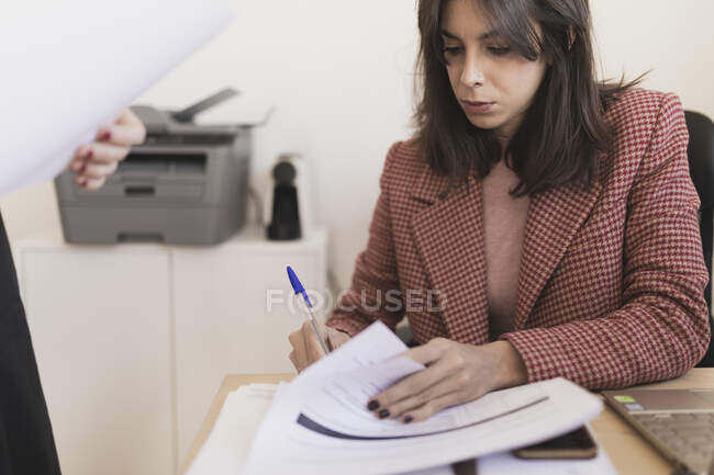 Crop femme debout près concentré jeune femme écriture dans les documents et assis à table avec ordinateur portable et smartphone dans le bureau — Photo de stock