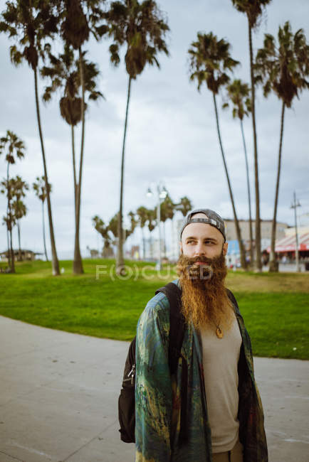 Вид збоку бородатого хлопця з рюкзаком, який дивиться геть, стоячи на тротуарі на вулиці сучасного міста — стокове фото