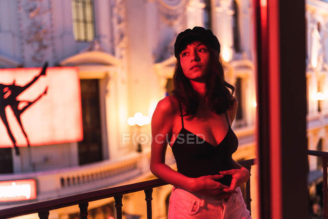 Young slim woman in cap standing near balcony in room in redness at night — Stock Photo