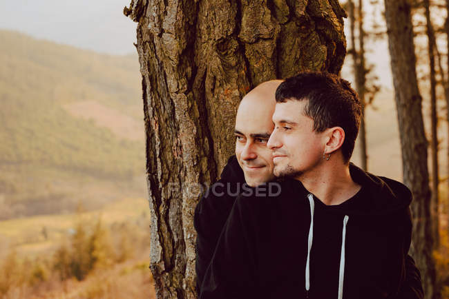 Dreamy homosexual couple embracing near tree in forest and picturesque view of valley — Stock Photo