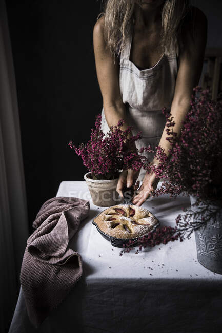 Mulher de colheita cozinheiro em avental de corte de bolo de ameixa na mesa com flores — Fotografia de Stock